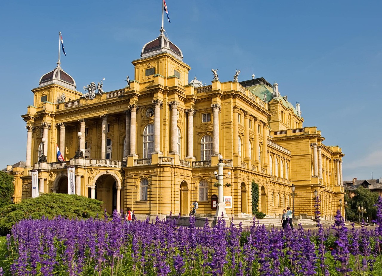 A yellow Baroque building with purple flowers in front of it 